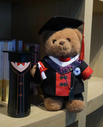 A mascot plush toy wearing a graduation gown sitting on a bookshelf as a campus keepsake